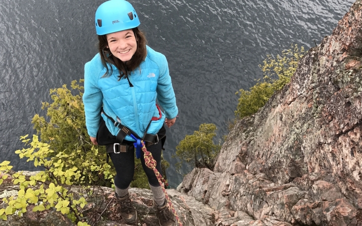 A young person wearing safety gear is secured by ropes as they stand on the edge of a cliff, high above a body of water.